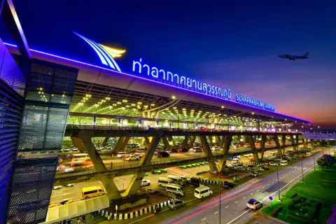 Der beleuchtete Flughafen Bangkok Suvarnabhumi bei Nacht, mit Autos auf mehrstöckigen Zufahrtsstraßen und einem Flugzeug am Himmel.