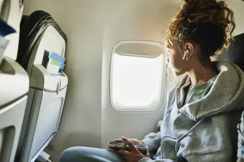 Junge Frau blickt aus dem Flugzeugfenster, vertieft in ihr Handy. Sie fragt sich, wie ist es zu fliegen, während sie die Wolken vorbeiziehen sieht.