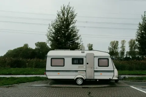 Ein weißer b96 wohnwagen steht auf einem gepflasterten Parkplatz, umgeben von grüner Wiese und Bäumen unter einem grauen Himmel.