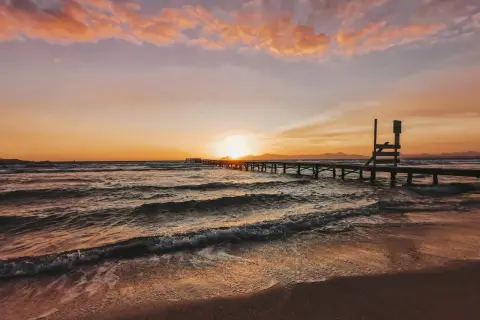 Sonnenuntergang über dem Meer an der Playa de Muro. Wellen brechen sanft am Strand, ein langer Holzsteg führt ins Wasser.