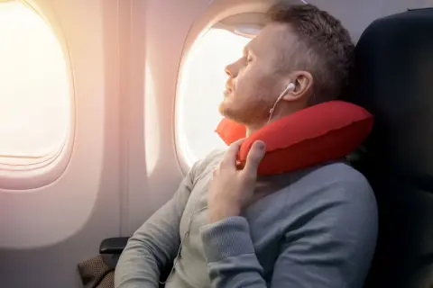 Mann schläft im Flugzeug mit rotem Nackenkissen. Ein Nackenkissen Flugzeug ist sinnvoll für bequemen Schlaf.