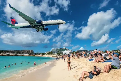 Ein Delta-Flugzeug landet tief über einem belebten Strand in der Karibik. Urlauber am **Karibik Flughafen** genießen die Sonne, während das Flugzeug über ihnen kreist.