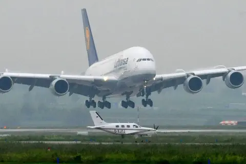 Ein riesiger A380 von Lufthansa landet, sein enormes Gewicht wird durch die vier Triebwerke und das Fahrwerk sichtbar. Ein kleines Propellerflugzeug wartet daneben.