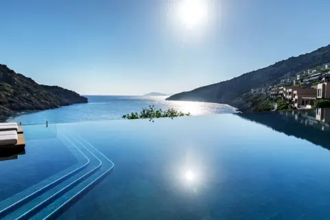 Infinity-Pool mit Blick auf die Bucht von Daios. Sonnenschein spiegelt sich im Wasser, Häuser schmiegen sich an den Hang.