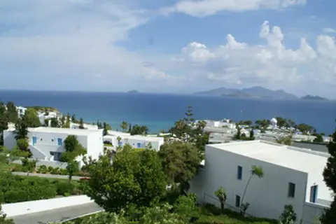 Blick auf das Robinson Daidalos Resort mit weißen Gebäuden und blauen Fenstern, umgeben von grüner Vegetation, am Meer mit Inseln am Horizont.