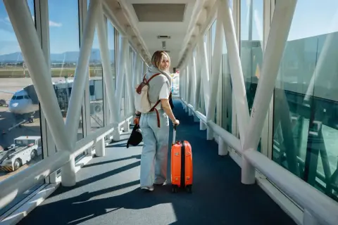 Junge Frau mit orangefarbenem Koffer und Rucksack geht durch einen Flugsteig. Ihr Flug mit Zwischenstopp beginnt.