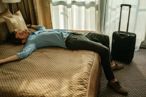 Erschöpfter Mann liegt auf einem Hotelbett, Koffer daneben. Der Jetlag von Asien nach Deutschland hat ihn völlig ausgeknockt.