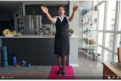 Eine Stewardess oder Flugbegleiterin in Uniform steht auf einer rosa Matte und macht Übungen. Im Hintergrund eine Küche und ein großes Fenster.