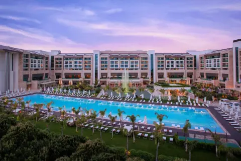 Ein luxuriöses Resort mit einem riesigen Pool und Palmen, das an das **Regnum Carya** erinnert. Abenddämmerung mit rosa Himmel.