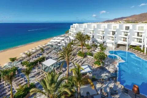 Traumhafter Blick auf das Riu Palace Jandia: Weißes Hotel, Pool, Palmen und der goldene Strand am türkisfarbenen Meer.