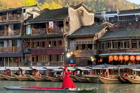 Ein Urlaub in China: Frau in rotem Kleid auf Boot vor traditionellen Häusern und Booten am Fluss.
