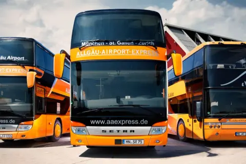 Drei orange-schwarze Doppeldeckerbusse von Allgäu Airport Express stehen bereit. Informieren Sie sich über die alex airport shuttle preise für Ihre Reise.