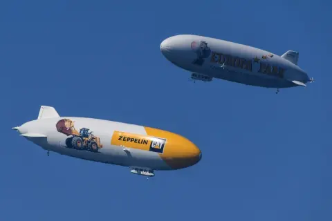 Zwei Zeppeline, einer mit einem Bagger und "Zeppelin CAT", der andere mit "Europa Park" und einem Koala, schweben am blauen Himmel.