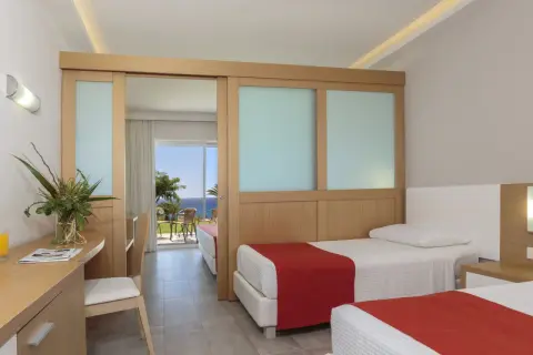Zwei Betten mit roten Decken im Rodos Princess Beach Hotel. Ein Schreibtisch mit Blumen und Blick aufs Meer.