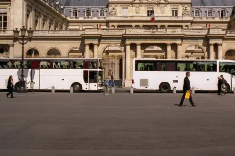 Weiße Reisebusse vor einem prächtigen Gebäude. Eine Frau geht vorbei. Wie kommt man am besten nach Paris? Mit dem Bus!