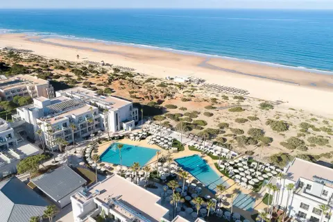 Luftaufnahme des Hipotels Barrosa Park mit Pools, Liegestühlen und Palmen, daneben ein breiter Sandstrand und das blaue Meer.