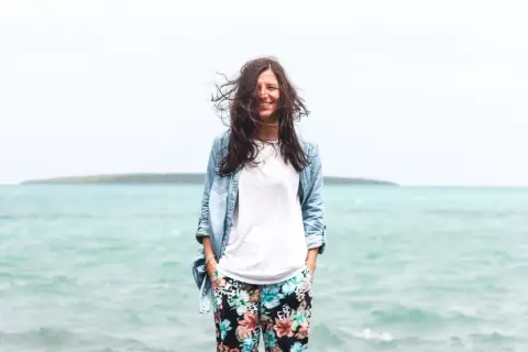 Frau mit wehendem Haar am Meer, die lächelt. Perfekt für die Packliste Mauritius: leichte Kleidung, Jeansjacke und bunte Hose.