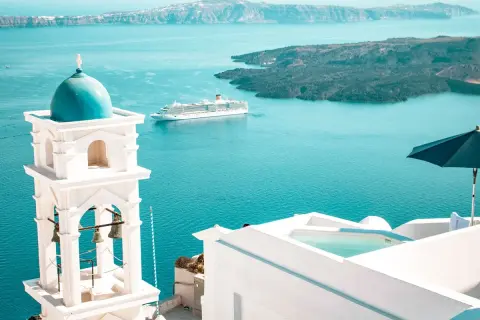 Traumhafter Urlaub Griechenland: Weißer Glockenturm mit blauer Kuppel, Kreuzfahrtschiff auf dem Meer, Infinity-Pool und Sonnenschirm.