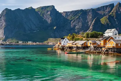 Idyllische Fischerhäuser auf Stelzen am kristallklaren Wasser vor majestätischen Bergen. Ein perfekter Urlaub in Norwegen.
