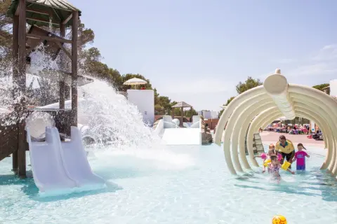 Kinder planschen im Wasserpark, während ein Wasserfall vom Rutschenturm herabstürzt. Ein Mann mit gelbem Shirt beaufsichtigt die Kleinen unter einem knochenartigen Tunnel in den Calimera Fido Gardens.