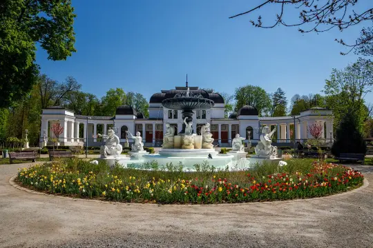 Prachtvoller Brunnen mit Skulpturen vor einem eleganten Gebäude in Cluj Napoca. Bunte Blumenbeete säumen den Weg.