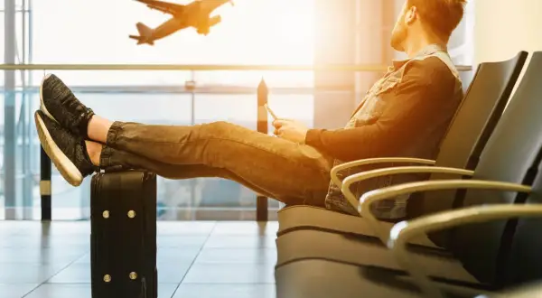 Mann mit Handy und Koffer wartet am Flughafen. Er überlegt, was kann man im Flugzeug machen, während ein Flugzeug startet.