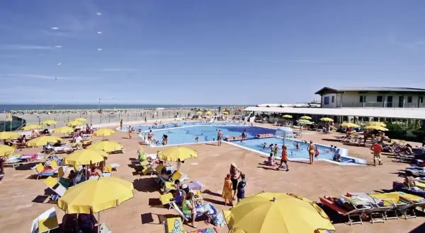 Sonnige Poolanlage mit vielen Liegen und gelben Sonnenschirmen am Rosapineta Sud. Kinder planschen im Wasser, während Möwen über dem Meer kreisen.