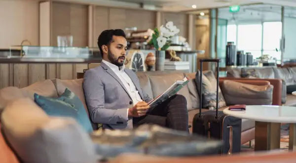 Mann im Anzug liest Zeitschrift in der airport VIP lounge. Sein Koffer steht bereit.