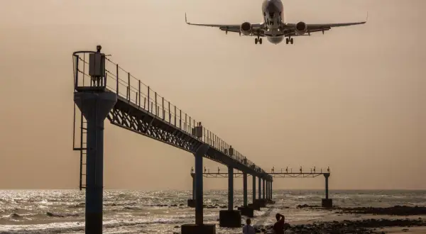 Zwei Personen genießen das Plane Spotting am Strand, während ein Flugzeug über eine Seebrücke hinwegfliegt.