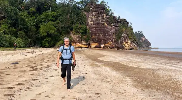 Mann mit Rucksack und Schuhen in der Hand geht barfuß am Strand entlang. Die Packliste für Malaysia ist komplett, die Natur ist üppig.