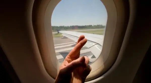 Hand mit überkreuzten Fingern vor Flugzeugfenster. Ein Zeichen der Hoffnung für alle Flugzeug-Passagiere.