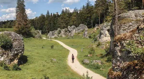 Frau wandert auf einem Kiesweg durch eine felsige Landschaft mit grünen Wiesen und Bäumen. Perfekt für einen entspannten outdoor urlaub.