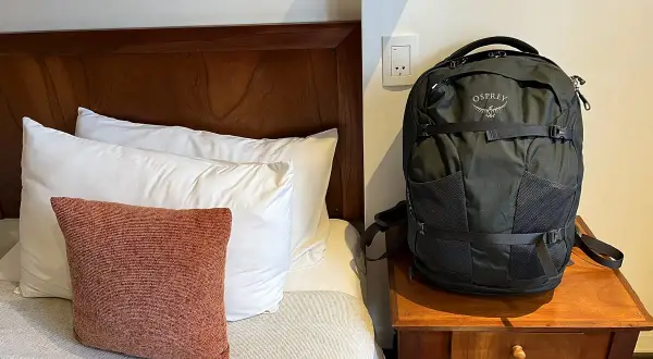 Schwarzer Osprey-Rucksack neben einem Bett mit Kissen. Perfekt für deine Packliste Mexiko.