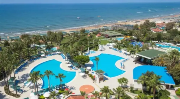 Blauer Poolbereich mit Palmen und Liegen, daneben ein langer Sandstrand mit vielen Sonnenschirmen und dem Meer. Ein Paradies am Strand.