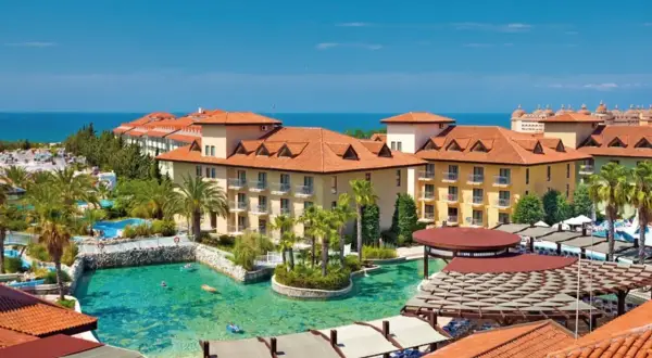 Ein luxuriöser Urlaub im Club Grand Aqua: Sonnige Gebäude mit roten Ziegeldächern, Palmen und ein türkisfarbener Pool mit Badegästen.