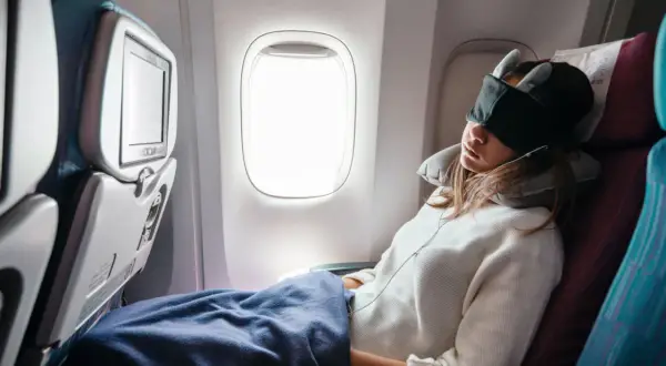 Frau schläft im Flugzeug, mit Schlafmaske und Nackenkissen, eingewickelt in eine Decke.