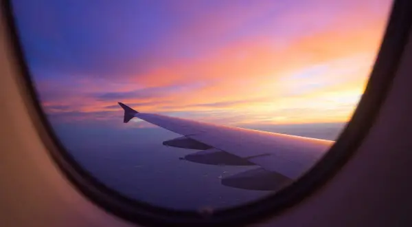 Flugzeugfenster mit Flügel und einem atemberaubenden Sonnenuntergang in Violett- und Orangetönen. Perfekt, um einen **stopover flug buchen** zu planen.