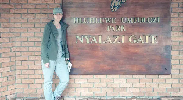 Frau am Eingangstor des Hluhluwe Umfolozi Parks, bereit für ihr Safari-Abenteuer. Ihre Kleidung ist perfekt für die Packliste.