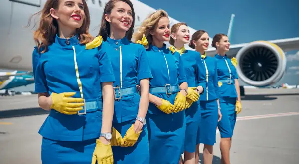 Fünf Flugbegleiterinnen in blauen Uniformen und gelben Handschuhen stehen vor einem Flugzeug. Sie sind bereit, ihre Passagiere zu begrüßen, denn das ist, was man als Flugbegleiterin macht.