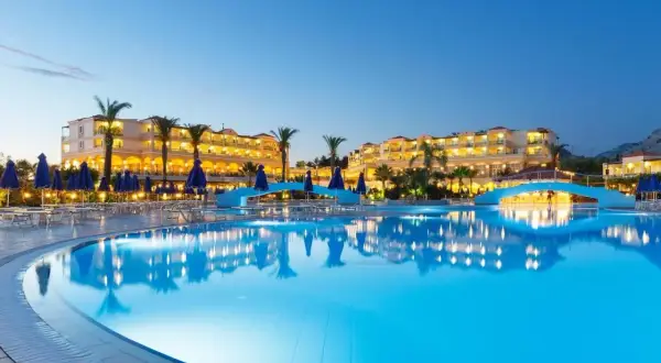 Ein beleuchteter Pool spiegelt das Lindos Princess Beach Hotel wider. Palmen und blaue Sonnenschirme säumen den Poolbereich.