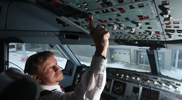Pilot im Cockpit prüft die Anzeigen. Er bedient einen Schalter, während draußen Schnee liegt.