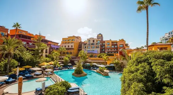 Sonnige Poolanlage mit bunten Gebäuden und Palmen im Riu Palace Teneriffa. Entspannung pur unter blauem Himmel.