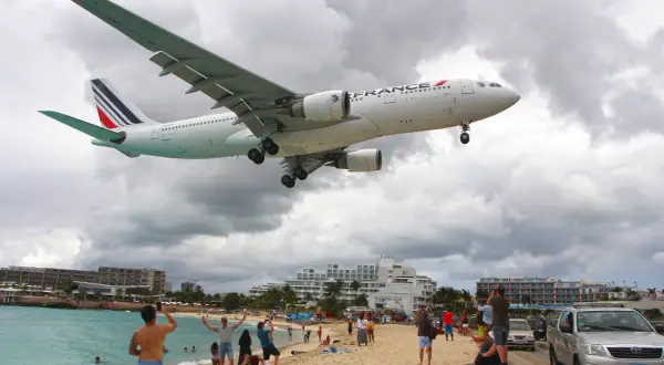 Air France Flugzeug landet auf einem Flughafen direkt am Strand. Menschen amüsieren sich am Meer, während das Flugzeug über sie hinwegfliegt.