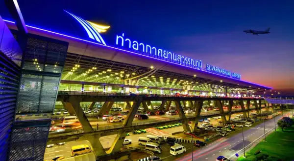 Der beleuchtete Flughafen Bangkok Suvarnabhumi bei Nacht, mit Autos auf mehrstöckigen Zufahrtsstraßen und einem Flugzeug am Himmel.