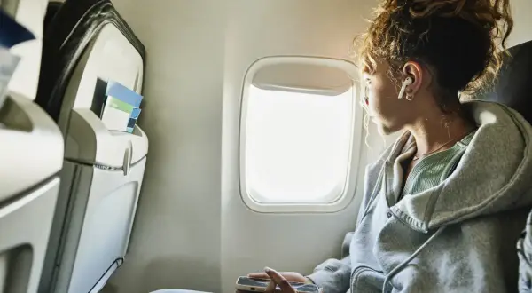 Junge Frau blickt aus dem Flugzeugfenster, vertieft in ihr Handy. Sie fragt sich, wie ist es zu fliegen, während sie die Wolken vorbeiziehen sieht.