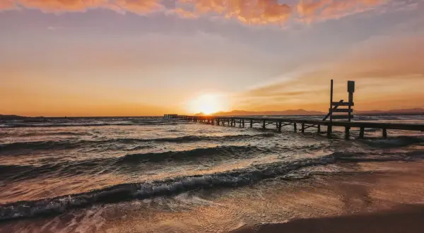 Sonnenuntergang über dem Meer an der Playa de Muro. Wellen brechen sanft am Strand, ein langer Holzsteg führt ins Wasser.