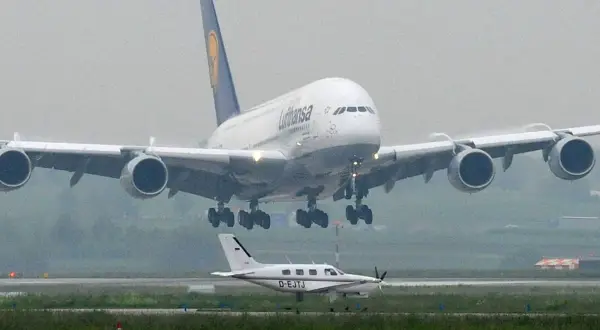 Ein riesiger A380 von Lufthansa landet, sein enormes Gewicht wird durch die vier Triebwerke und das Fahrwerk sichtbar. Ein kleines Propellerflugzeug wartet daneben.
