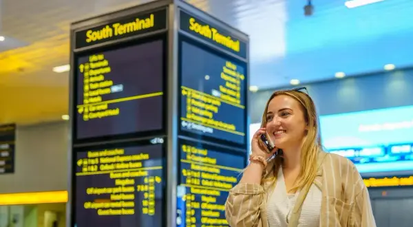 Frau telefoniert am London Flughafen vor einer Anzeigetafel für den South Terminal.