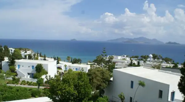 Blick auf das Robinson Daidalos Resort mit weißen Gebäuden und blauen Fenstern, umgeben von grüner Vegetation, am Meer mit Inseln am Horizont.