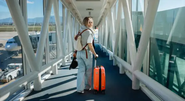 Junge Frau mit orangefarbenem Koffer und Rucksack geht durch einen Flugsteig. Ihr Flug mit Zwischenstopp beginnt.